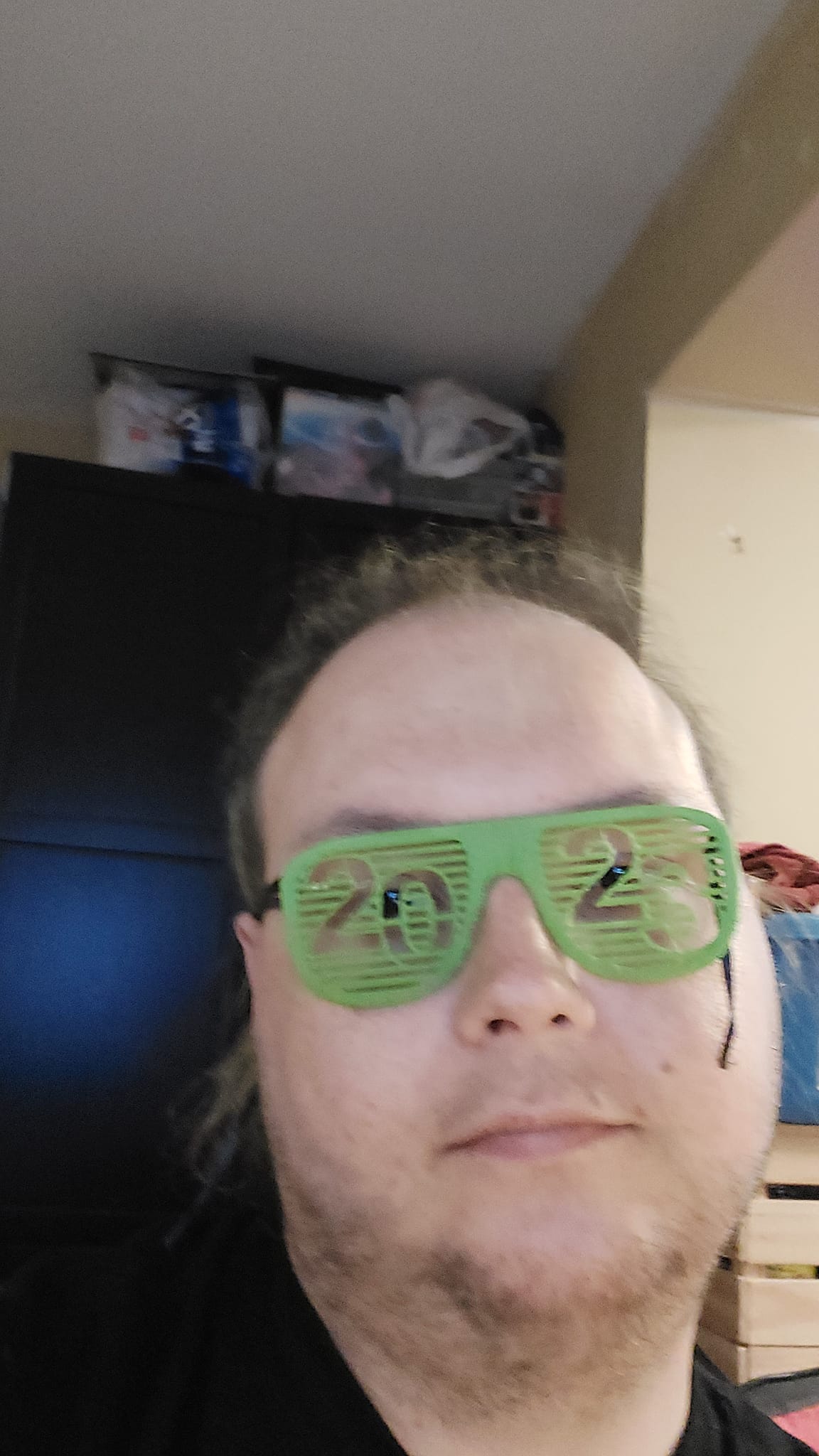 A man sits in a computer chair, showing off green plastic glasses that have 20 and 23 written on the lenses